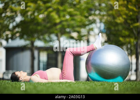 Giovane donna incinta giacente sul tappeto di esercizio su erba verde ha sollevato il suo piede sulla palla e fare gli esercizi nel parco Foto Stock