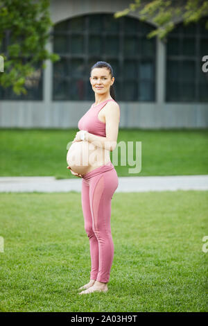 Ritratto di bella donna incinta in rosa abbigliamento sportivo in piedi e toccando il suo ventre mentre la formazione nel parco all'aperto Foto Stock