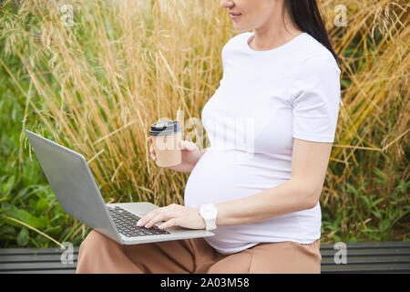 Close-up della donna incinta seduta sul banco di lavoro con computer portatile sulle ginocchia di lavoro e di bere il caffè all'aperto Foto Stock
