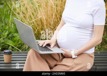 Close-up della donna incinta in linea di lavoro sul computer portatile mentre è seduto sul banco di prova all'aperto Foto Stock
