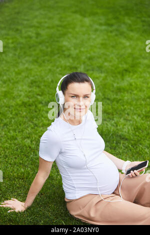 Ritratto di giovane donna incinta in cuffia seduti su erba verde e sorridente in telecamera mentre si ascolta la musica di relax sul suo telefono cellulare Foto Stock
