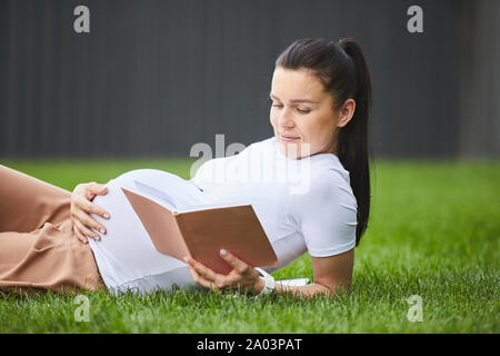 Giovane donna incinta con scuri capelli lunghi giacente su erba verde e rilassante durante la lettura di un libro Foto Stock