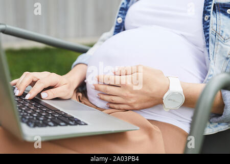 Close-up della donna incinta di toccare la sua pancia mentre è seduto e utilizzando il computer portatile all'aperto Foto Stock