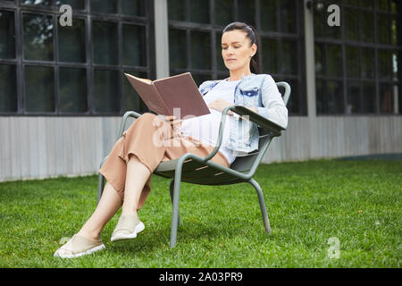 Giovane donna incinta in appoggio su una sedia e la lettura di un libro interessante circa la gravidanza su aria fresca Foto Stock