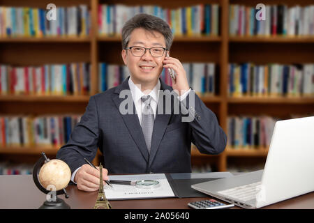 Un ritratto di un asiatico di mezza età imprenditore maschio seduti ad una scrivania, sorridente e parlare al telefono. Foto Stock