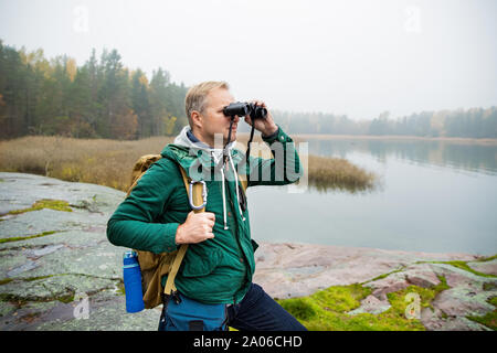 Uomo maturo ad esplorare la Finlandia in autunno, cercando nella nebbia attraverso il binocolo. Escursionista con grande zaino in piedi sulla roccia di muschio. Paesaggio scandinavo Foto Stock