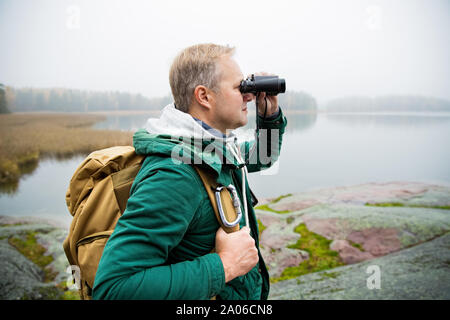 Uomo maturo ad esplorare la Finlandia in autunno, cercando nella nebbia attraverso il binocolo. Escursionista con grande zaino in piedi sulla roccia di muschio. Paesaggio scandinavo Foto Stock