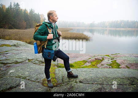 Uomo maturo ad esplorare la Finlandia in autunno, cercando nella nebbia. Escursionista con grande zaino in piedi sulla roccia di muschio. Paesaggio scandinavo con misty e mare Foto Stock