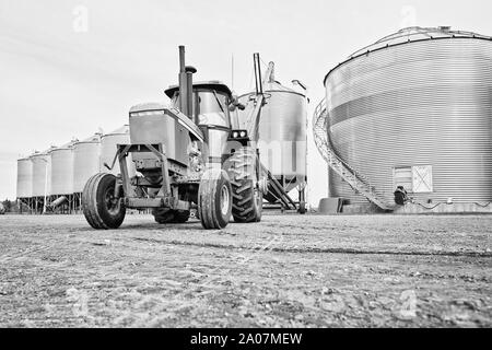 Un trattore con attaccato un backup della coclea fino a una fila di acciaio inossidabile contenitori del cereale in un'agricoltura farm yard in un paesaggio autunnale Foto Stock