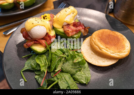 Avocado benedetto. Uovo affogato servita su una metà di avocado con salsa olandese e bacon, pane bun e insalata verde su un lato Foto Stock