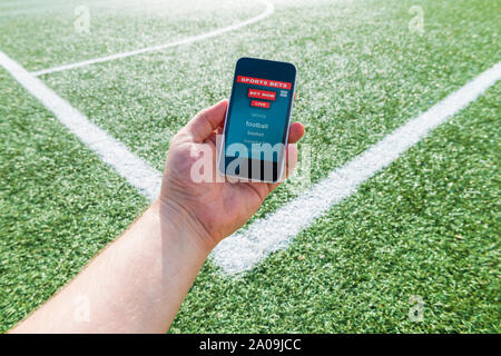 Una mano d'uomo in possesso di un telefono mobile in cui la interfaccia di scommesse sportive pagina è visto, sullo sfondo verde di un campo da calcio. Scommesse online Foto Stock