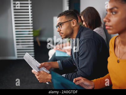 Vista laterale di un fiducioso giovane americano africano uomo che indossa occhiali azienda riprendere in mano in attesa per il colloquio di lavoro con altri candidati Foto Stock