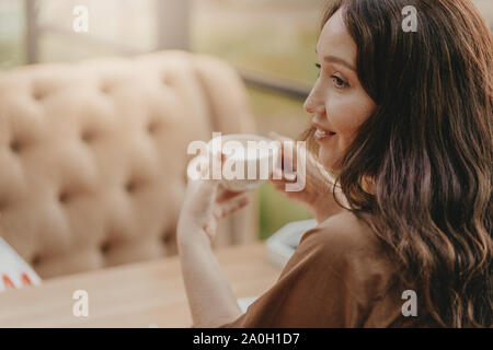 Affascinante brunette donna con lunghi capelli ricci seduta alla finestra nel cafe con la tazza di caffè nelle mani Foto Stock