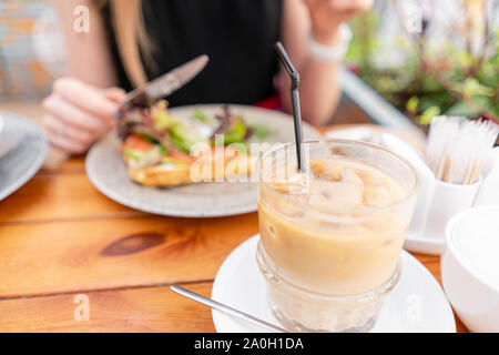 La messa a fuoco in primo piano sul caffè freddo con cubetti di ghiaccio. Una giovane donna è avere la prima colazione in una caffetteria estiva. Foto Stock