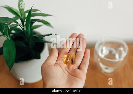 Integratori alimentari. Mano azienda omega 3 due capsule al di sopra di un tavolo di legno con un bicchiere di acqua. Mattina dose di vitamina. Olio di pesce in pillole. Assistenza sanitaria Foto Stock