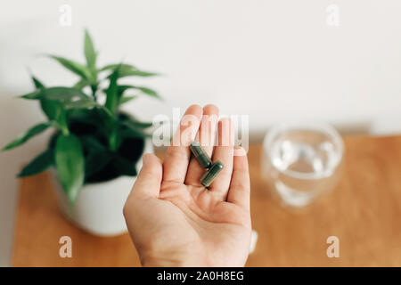 Mano che tiene la spirulina capsule al di sopra di un bicchiere di acqua su un tavolo di legno. Mattina vitamina pillola di nutrienti. Integratori alimentari. Assistenza sanitaria e di trattamento. Foto Stock