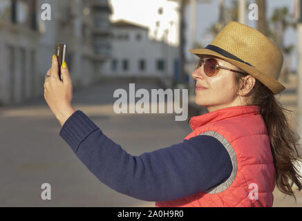 Donna attraente nel cappello e occhiali prende un selfie su una soleggiata giornata autunnale Foto Stock