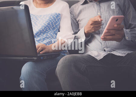 Donna al lavoro su laptop e uomo utilizzando il telefono cellulare entrambi sono seduti insieme sul divano di casa. Foto Stock