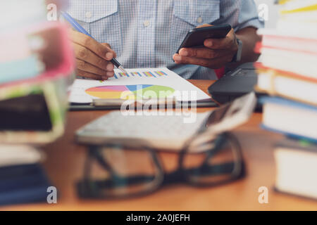 L uomo è utilizzando il telefono cellulare e lavorare con la pila di documenti sulla scrivania in ufficio, imprenditore è analisi di marketing con il grafico delle statistiche, Business e O Foto Stock