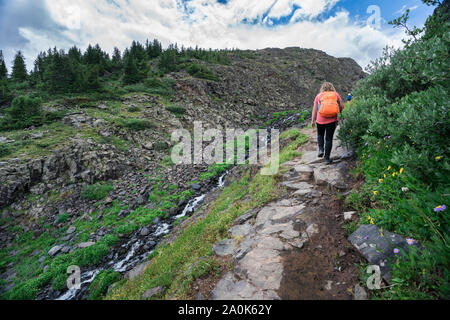 La bionda ragazza adolescente con zaino arancione passeggiate lungo un torrente di montagna su un sentiero roccioso in San Juan Mountains su un nuvoloso giorno di estate, Weminuche selvatica Foto Stock