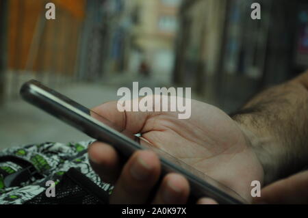 Man mano che tiene uno smartphone in strada e la città al di fuori della messa a fuoco in background-uomo seduto in una banca in strada con il suo cellulare Foto Stock