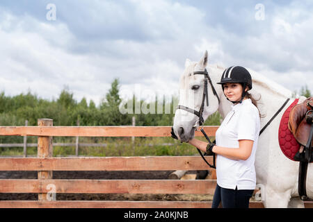 Giovane donna in abito equestre e delle corse ippiche bianco muovendosi lungo la recinzione di legno Foto Stock