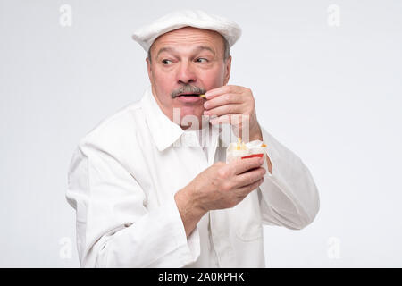 Funny affamato barbuto maschio chef cuoco di mangiare cibo spazzatura isolati su sfondo bianco alla ricerca geografica cauto. Studio shot Foto Stock
