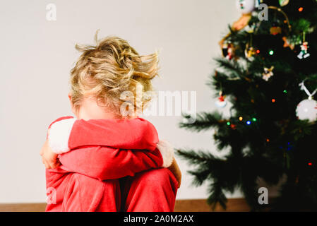 Bambino con travestimento, ansioso e triste il giorno di Natale con un volto arrabbiato accanto all'albero di Natale in attesa di Santa Foto Stock