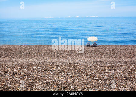 Due vuoto chaise longue e bianco ombrellone sulla bellissima lonely spiaggia ghiaiosa Foto Stock