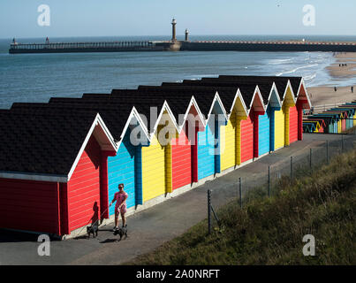 Una donna cammina i suoi cani passato dipinto luminosamente beach capanne in Whitby, nello Yorkshire, come le temperature sono attesi per volare a 26C in alcune parti del paese questo fine settimana. Foto Stock