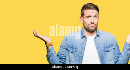 Uomo bello con gli occhi blu e la barba indossando giacca denim clueless e espressione confusa con le braccia e le mani alzate. Concetto di dubbio. Foto Stock