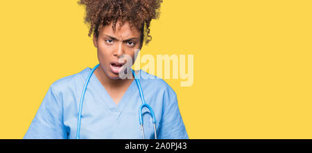 Giovane africano medico americano donna sullo sfondo isolato in shock viso, cercando scettici e sarcastico, stupisce con la bocca aperta Foto Stock