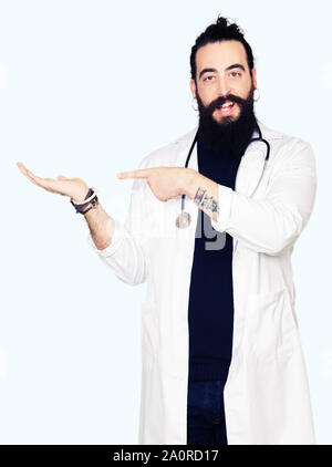 Medico con capelli lunghi che indossa medical e il cappotto di uno stetoscopio stupiti e sorridente alla fotocamera durante la presentazione con la mano e puntando con il dito. Foto Stock