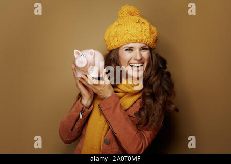 Felice donna alla moda in giallo berretto e sciarpa che mostra salvadanaio su sfondo di bronzo. Foto Stock