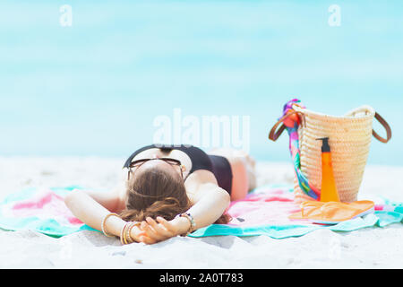 Elegante medioevo donna con lunghi capelli ricci in nero elegante costume da bagno su una spiaggia bianca dormendo mentre l'abbronzatura. Foto Stock