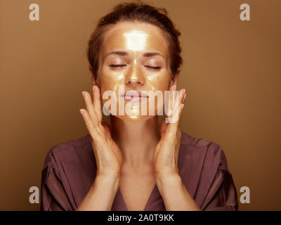 Ritratto di rilassata medioevo donna con golden cosmetici maschera viso rendendo il massaggio del viso isolato su sfondo beige. Foto Stock