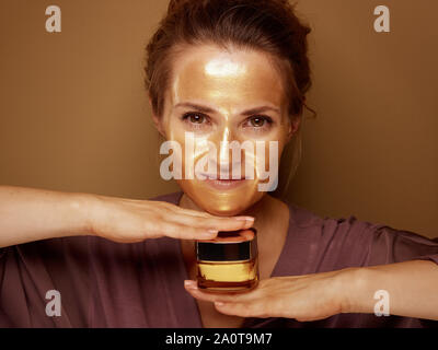 Ritratto della moderna casalinga con golden cosmetic maschera facciale di contenimento del prodotto cosmetico jar isolato su sfondo beige. Foto Stock