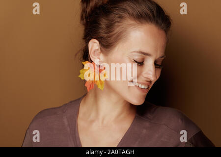 Ciao autunno. Sorridendo elegante 40 anno vecchia donna in accappatoio con foglie di autunno orecchini su sfondo marrone. Foto Stock