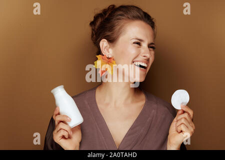 Ciao autunno. Sorridendo medioevo donna in un accappatoio con foglie di autunno orecchini azienda pulizia viso latte e tampone di cotone su sfondo marrone. Foto Stock