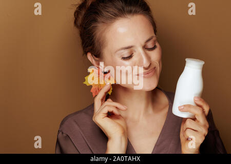 Ciao autunno. Felice elegante 40 anno vecchia donna in accappatoio con foglie di autunno orecchino e pulizia viso latte su sfondo di bronzo. Foto Stock