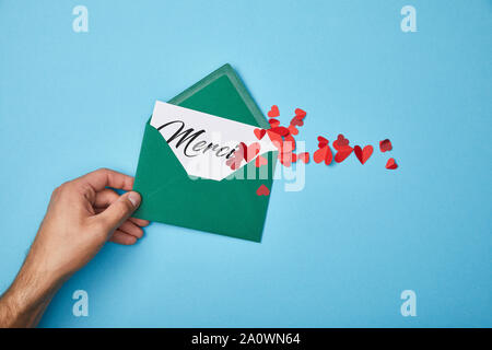 Vista ritagliata dell uomo con busta verde con merci di scritte su carta bianca e il taglio della carta cuori rossi su sfondo blu Foto Stock