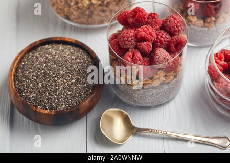 Occhiali con yogurt, fiocchi di avena e lamponi vicino al recipiente di legno con chia semi sul tavolo bianco Foto Stock