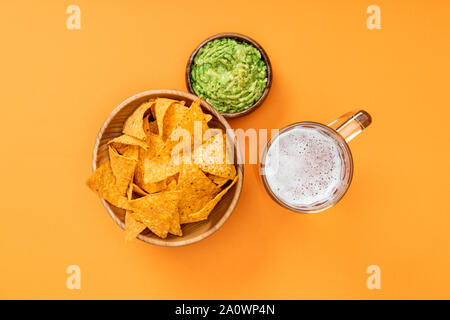 Vista dall'alto di croccante di nachos nella ciotola di legno vicino guacamole e birra su sfondo arancione, cucina messicana Foto Stock