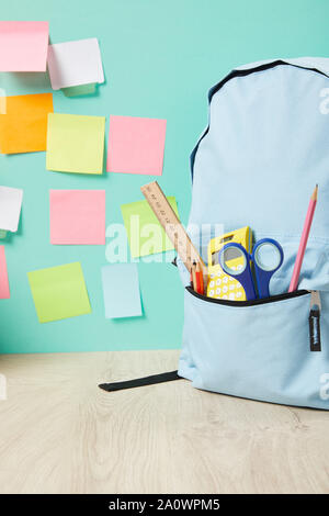 Scuola zaino blu con forniture in tasca vicino multicolore di sticky notes sulla parete turchese Foto Stock