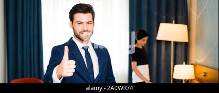 Panoramica di felice receptionist che mostra il pollice fino nei pressi di cameriera in camera di albergo Foto Stock