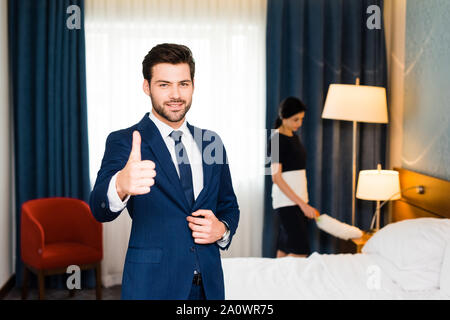 Messa a fuoco selettiva di felice receptionist che mostra il pollice fino nei pressi di cameriera in camera di albergo Foto Stock