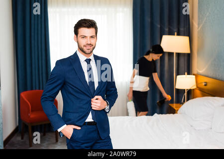 Felice receptionist in piedi con la mano in tasca vicino cameriera nella camera di albergo Foto Stock