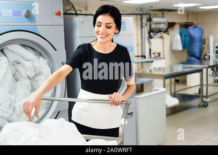 Messa a fuoco selettiva di sorridente cameriera vicino a cestello con white bed sheets Foto Stock