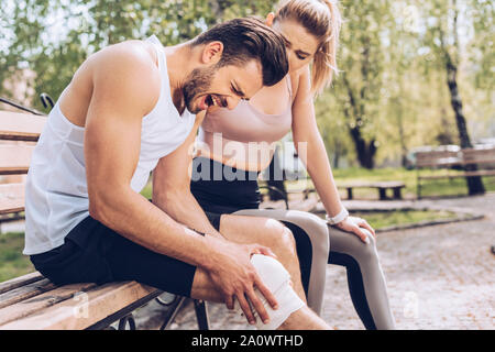 Giovane donna in sportswear seduta sul banco vicino a feriti sportivo soffre di dolori al ginocchio Foto Stock