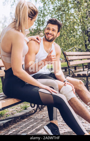 Feriti sportivo sorridente e mostrando un pollice alzato durante la seduta sul banco vicino sportive Foto Stock